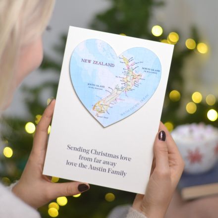 Pesonalised map heart card with personalised message printed underneath. White card with New Zealand map. Held in hands in front of Christmas lights.