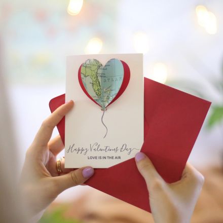 Valentine's card with map balloon in the shape of a heart with 'Happy Valentine's day - love is in the air' printed beneath. Card held against red envelope.
