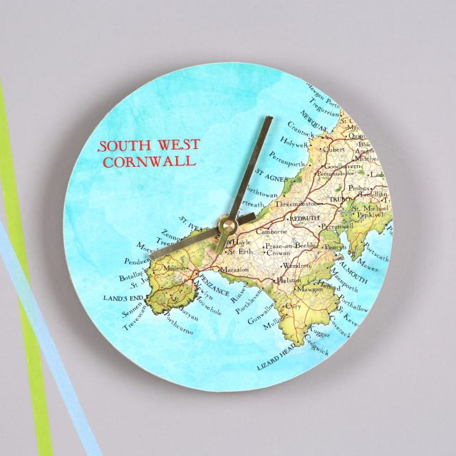 Round wall clock featuring map of Cornwall, hung on grey wall with gold hands.